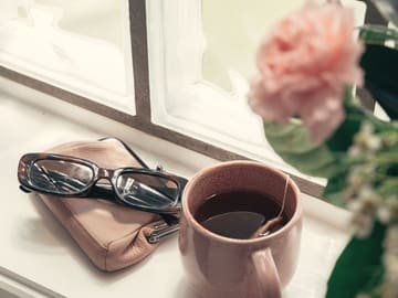 Lunettes posées près d’une tasse de thé, au bord d’une fenêtre fleurie